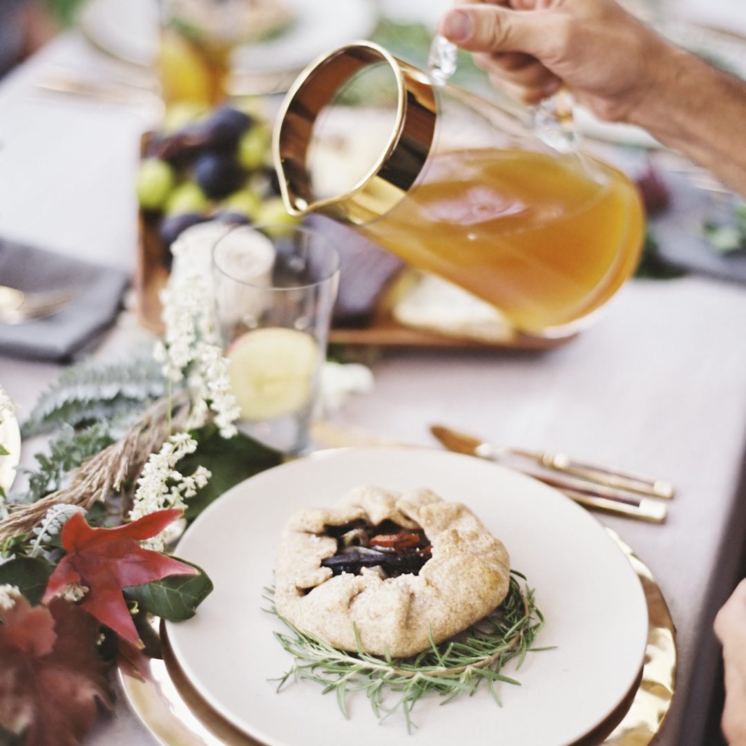 A group of people around a table in a garden. A celebration meal, with table settings and leafy decorations. A person pouring drinks into glasses.