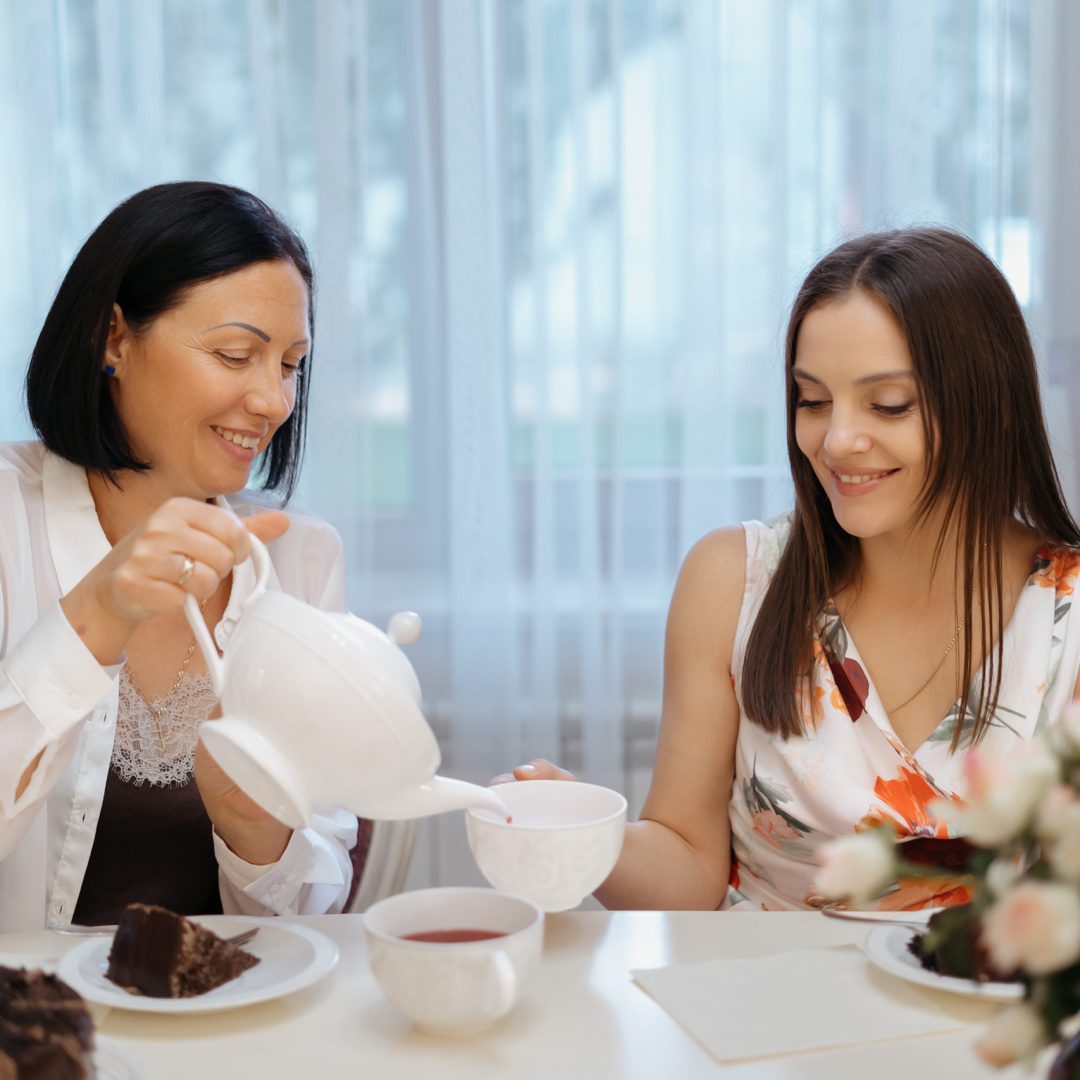 Woman pouring tea from a white teapot in white cup at home while sitting at the table and serving sisters smiling